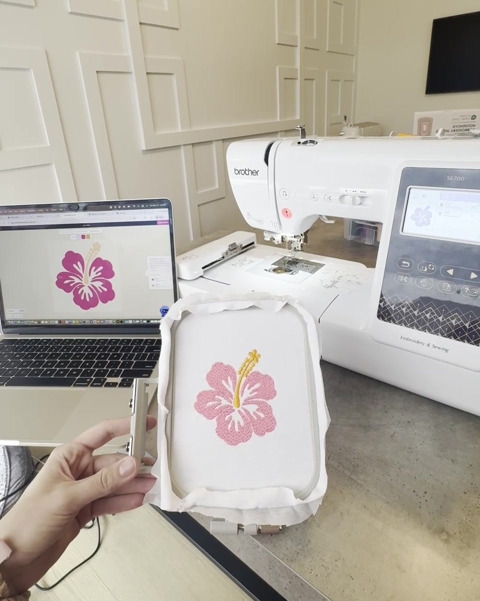 The same pink and yellow hibiscus stitched on white fabric in a hoop beside a Brother SE700 and the laptop running Trace.